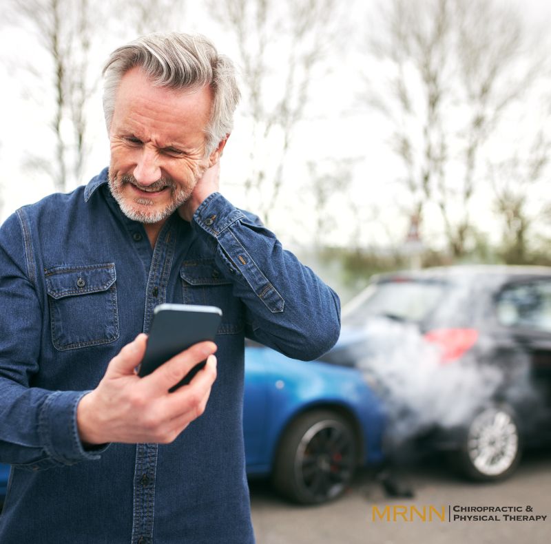 'n Man staan langs twee beskadigde motors, hou sy nek vas van pyn en kyk na sy foon, wat 'n onlangse motorongeluk suggereer. 'n Man staan langs twee beskadigde motors, hou sy nek vas van pyn en kyk na sy foon, wat 'n onlangse motorongeluk suggereer